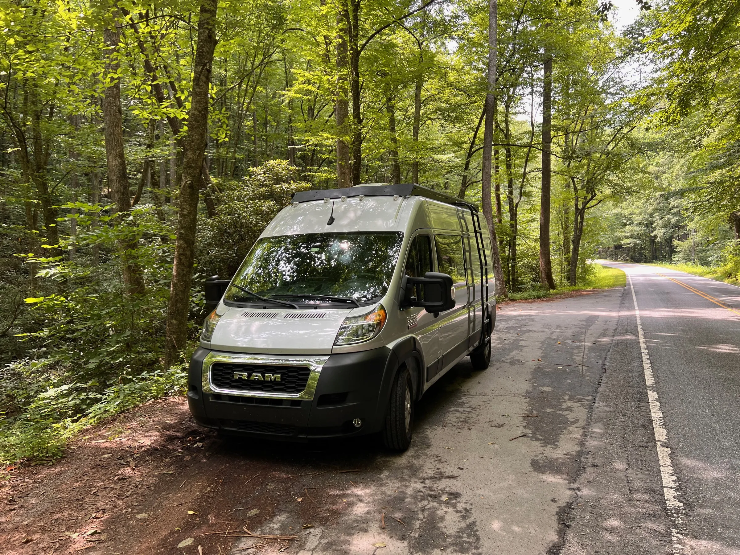 Silver 2022 Ram ProMaster 2500 high-roof van with black roof rack and side ladder parked on a forested mountain road