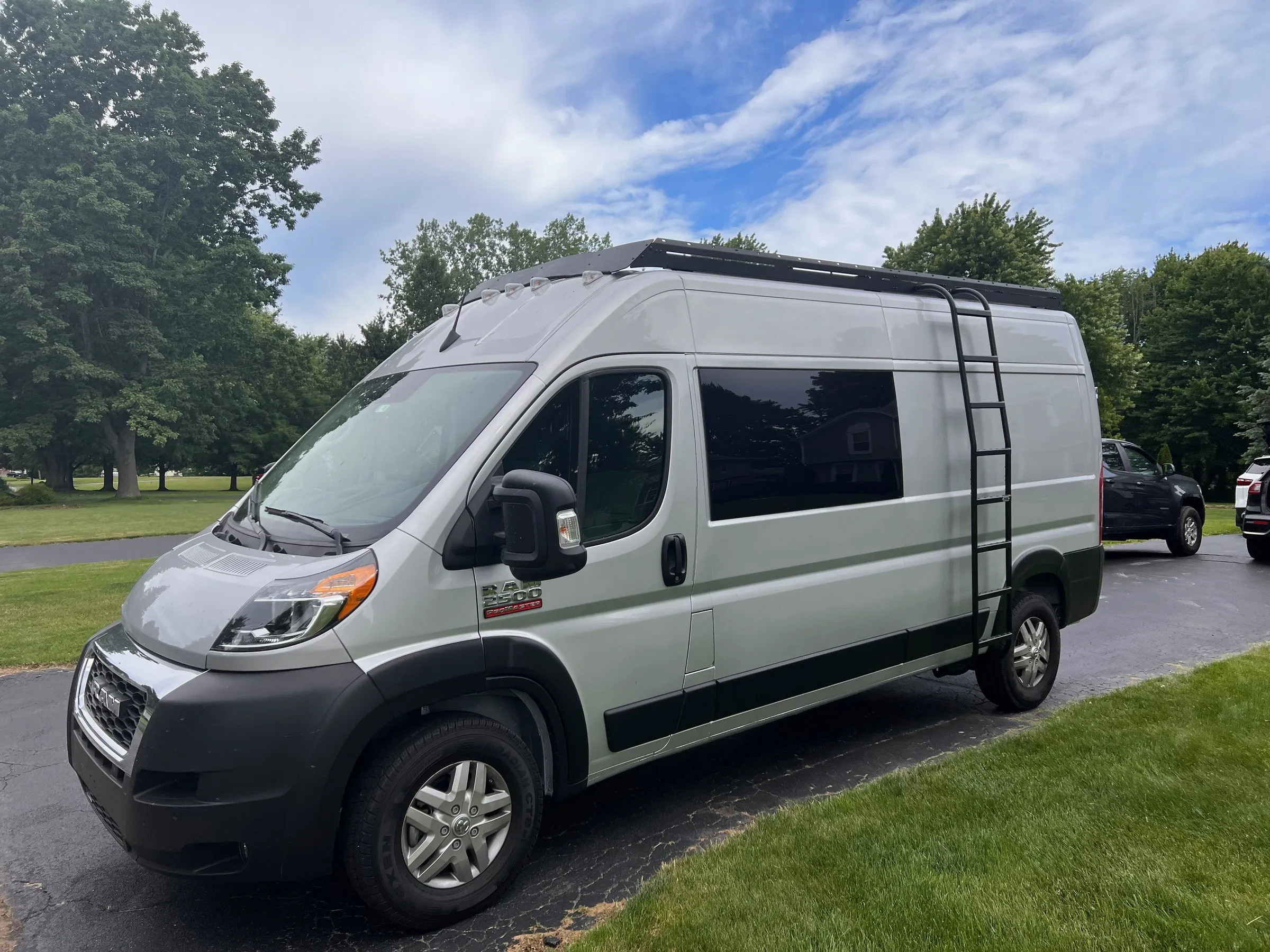 Passenger side of the silver ProMaster showing tinted side windows, full-length roof rack and rear ladder, parked on a driveway