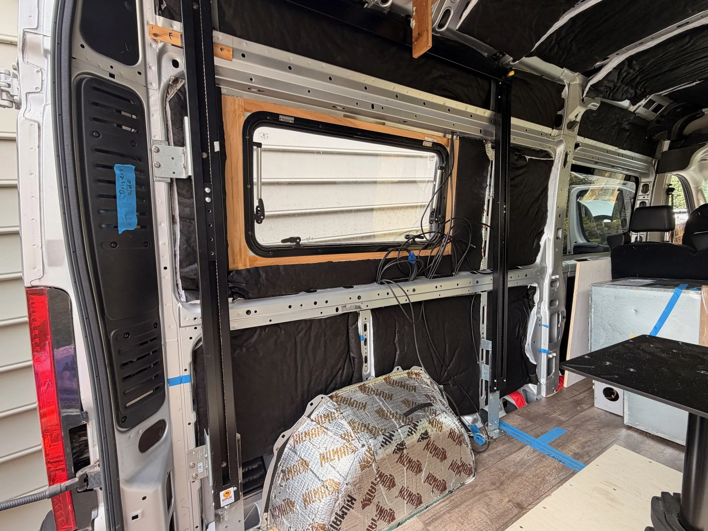 Driver-side rear wall with black insulation, an installed framed window, wiring runs and vertical bed-lift tracks
