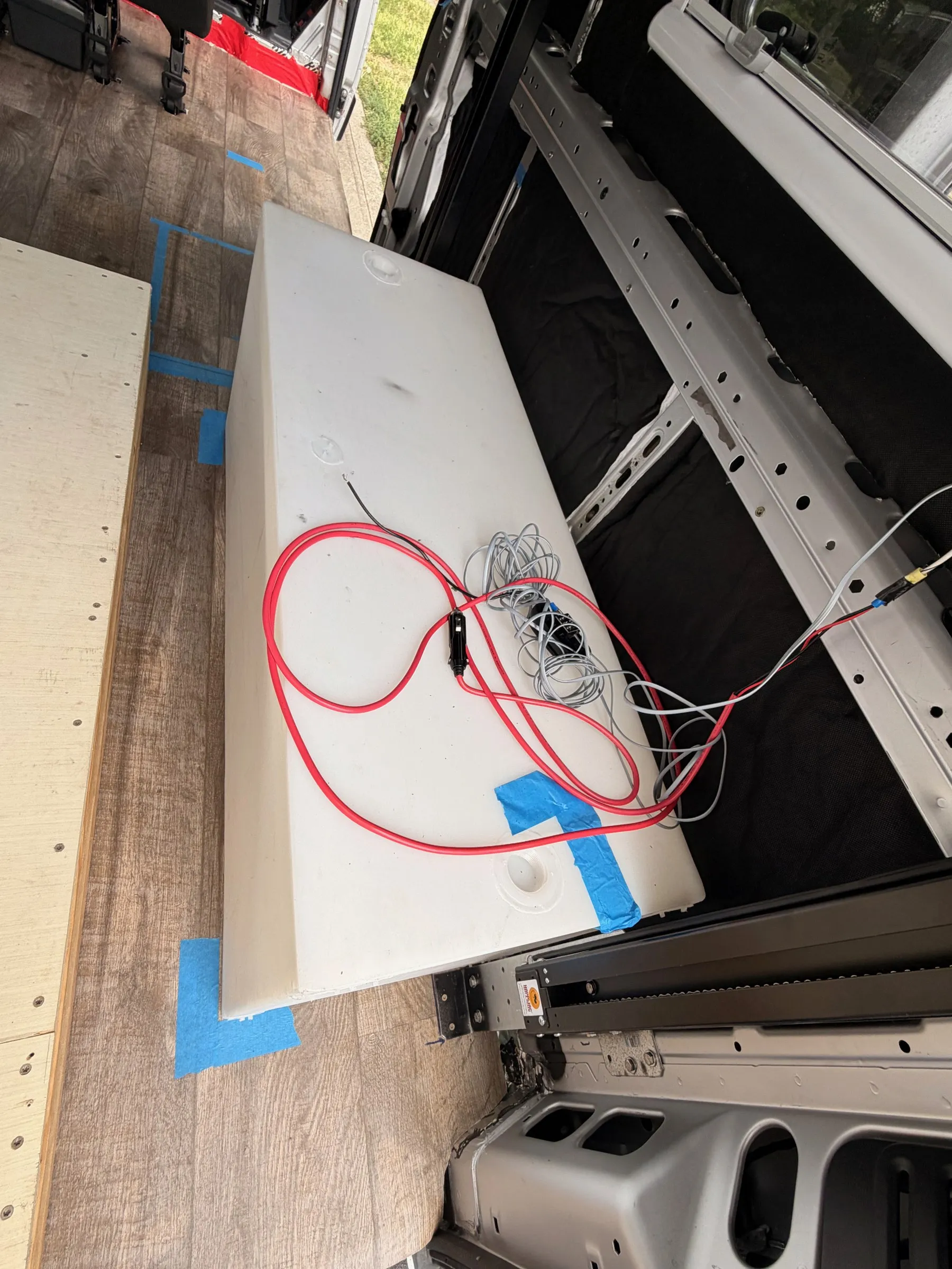 White rectangular poly water tank with wiring coiled on top, set along the passenger-side wall